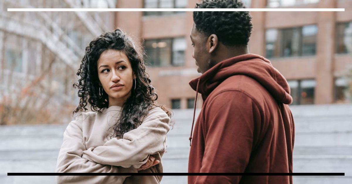 Duas pessoas conversam na rua; uma mulher de braços cruzados encara um homem, sugerindo desacordo.
