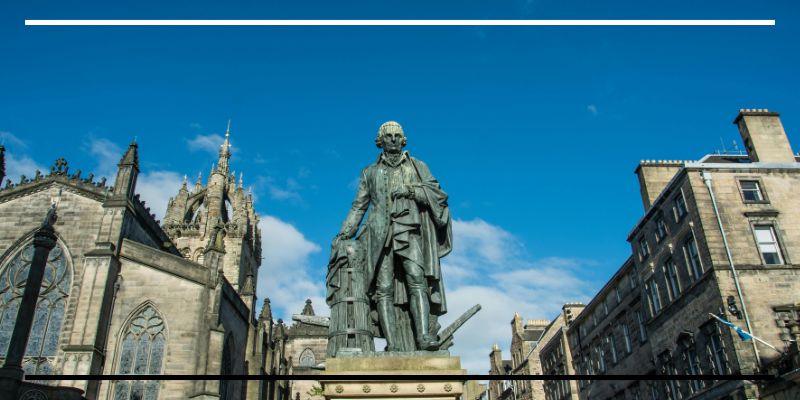 Estátua de Adam Smith em uma praça de Edimburgo, Escócia, com prédios históricos e céu azul ao fundo.
