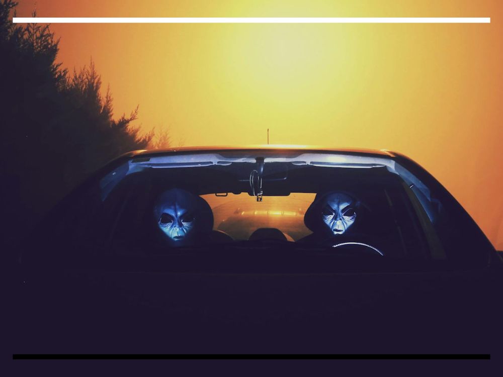 Dois alienígenas iluminados dentro de um carro à noite, com luz intensa ao fundo.