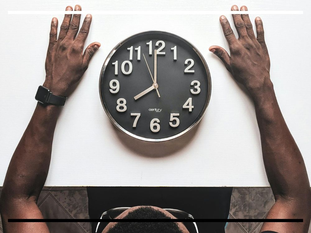 Homem olhando para um relógio sobre a mesa, simbolizando a relação entre tecnologia, tempo e rotina humana.
