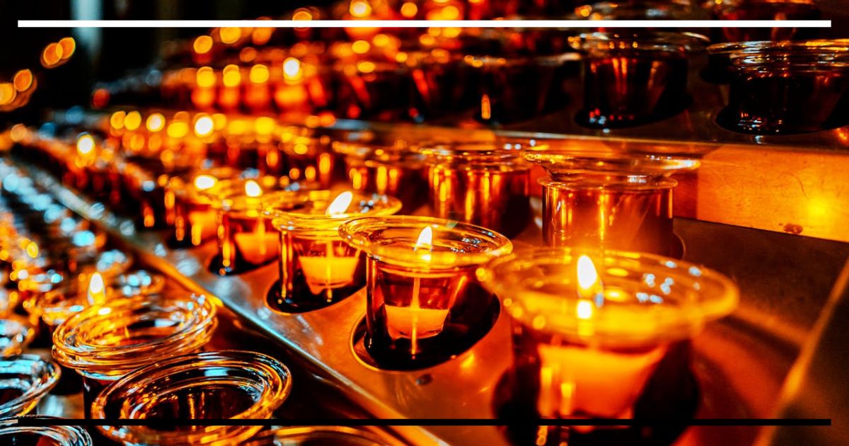 Velas acesas em fileiras iluminando um ambiente escuro, evocando presença e ausência simultâneas.