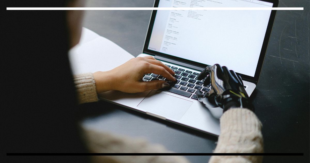 Close de teclado com uma mão orgânica e outra protética digitando — ambíguo se é um humano com mão de androide ou um androide com mão humana.