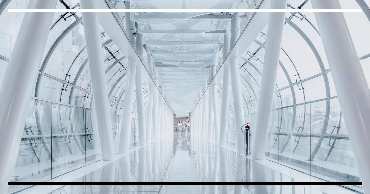 Corredor envidraçado, branco e simétrico, com pilares e paredes de vidro; piso reflexivo e duas pessoas ao fundo, tudo visível de ponta a ponta.
