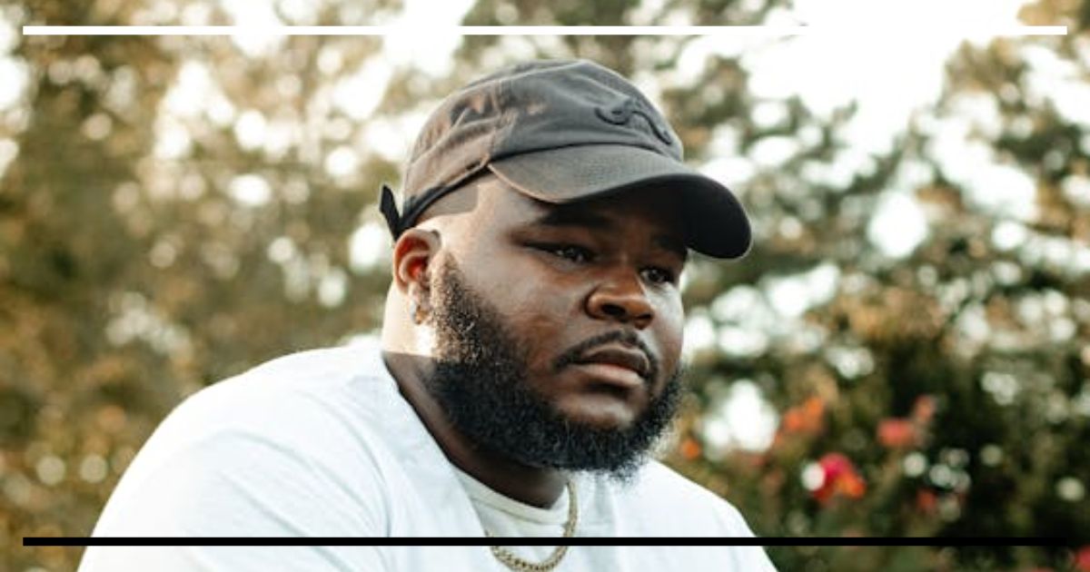Homem com boné preto e barba, expressão pensativa ao ar livre, luz suave ao entardecer.