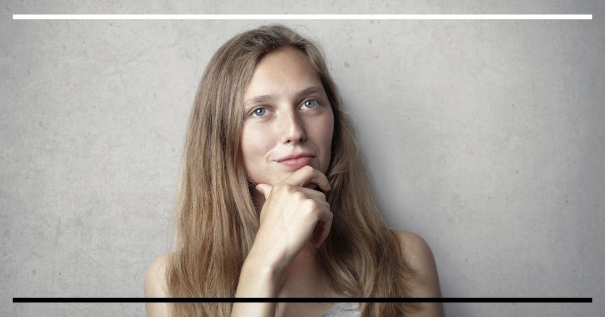 Mulher jovem, olhar pensativo, mão no queixo, fundo de parede cinza