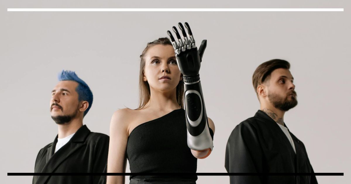 Três pessoas em estúdio neutro, com estética futurista: mulher ao centro com braço biônico erguido; dois homens ao fundo usando próteses de perna e roupas pretas minimalistas.