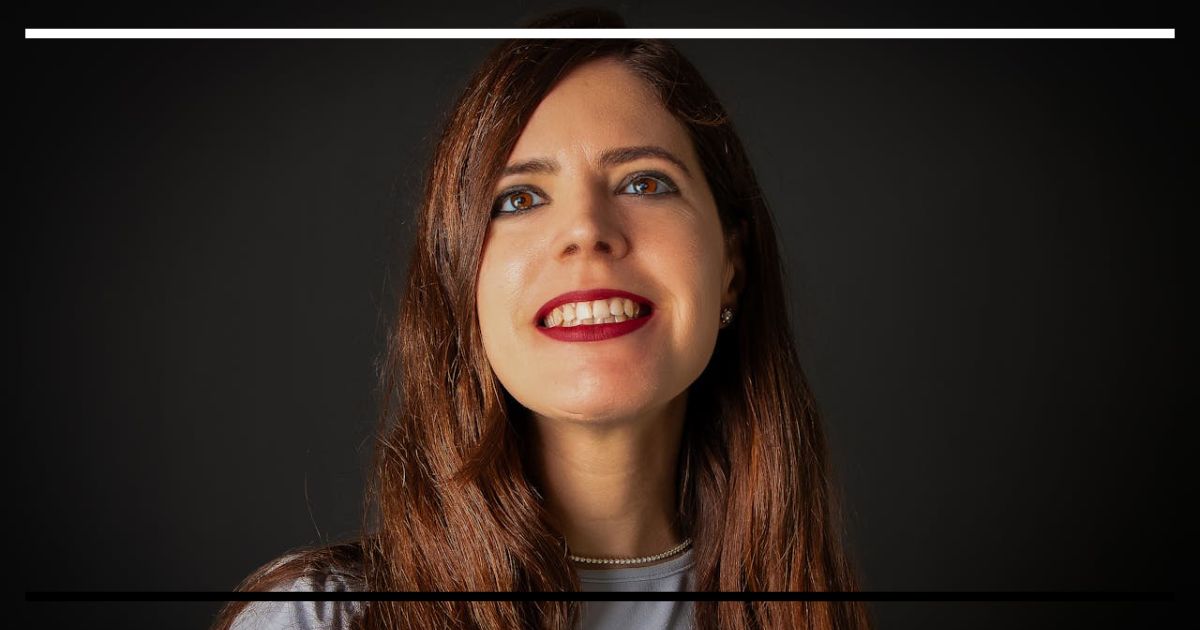 Retrato em close de uma mulher com cabelo castanho e sorriso amplo, vestindo blusa cinza, sobre fundo escuro.