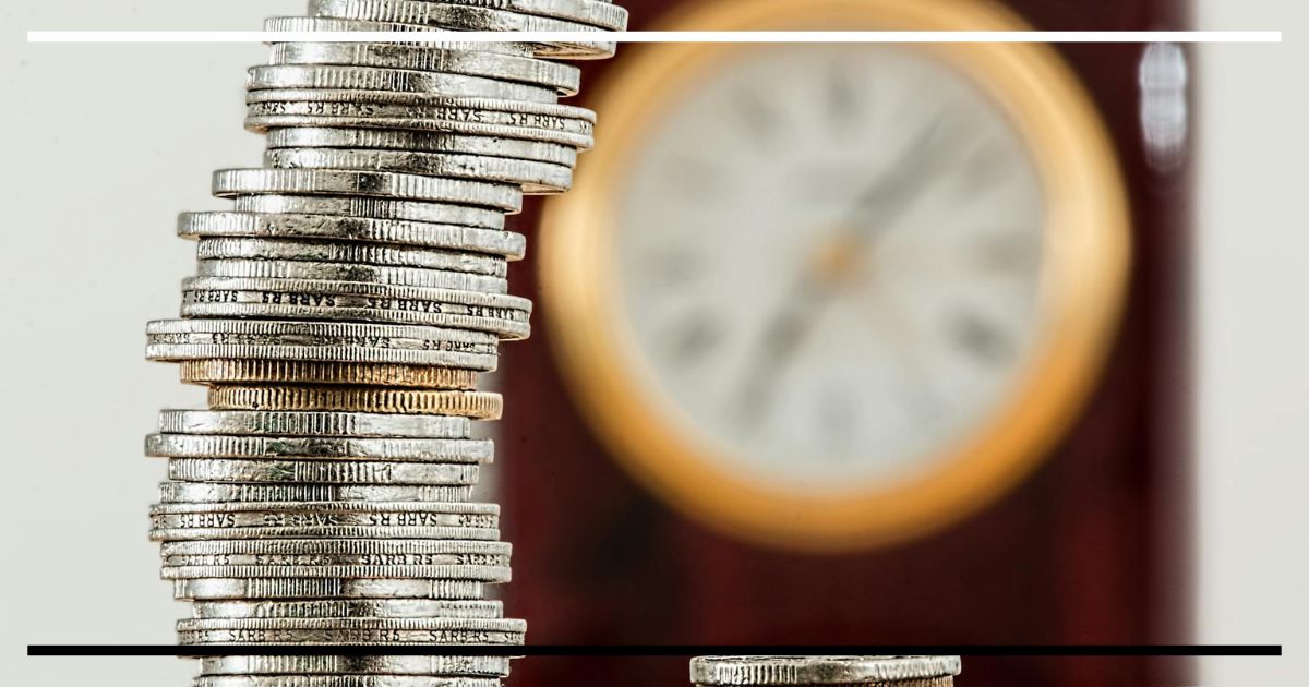 Pilha de moedas em close com relógio desfocado ao fundo, representando o preço do tempo e a taxa de juros de equilíbrio