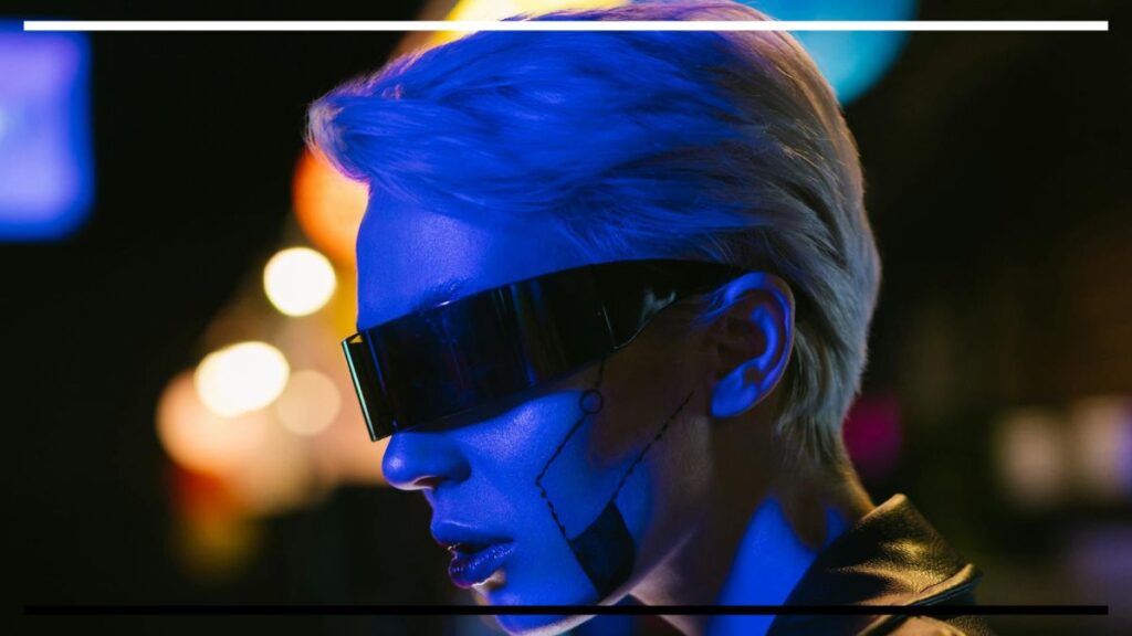 Close de perfil de pessoa com cabelo curto e claro, usando visor preto largo que cobre os olhos; rosto pintado com linhas e círculos que sugerem implantes/biomodificação.