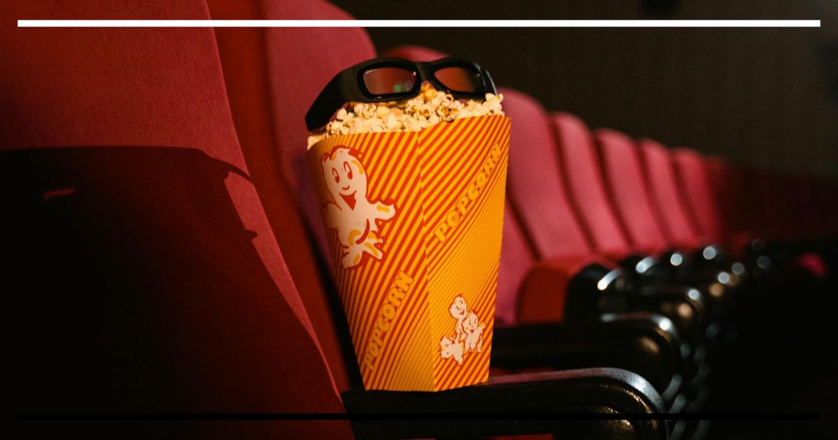 Balde de pipoca com óculos 3D sobre a poltrona em sala de cinema; fileiras vermelhas ao fundo.