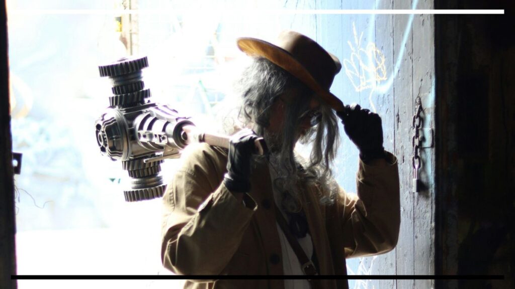 Pessoa de chapéu marrom, cabelo grisalho e óculos, com luvas pretas, tocando a aba do chapéu; sobre o ombro, um grande mecanismo metálico com engrenagens.
