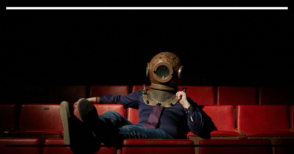 Pessoa sentada em poltrona vermelha de cinema usando capacete antigo de mergulho; sala escura ao fundo, clima de humor e estranhamento.