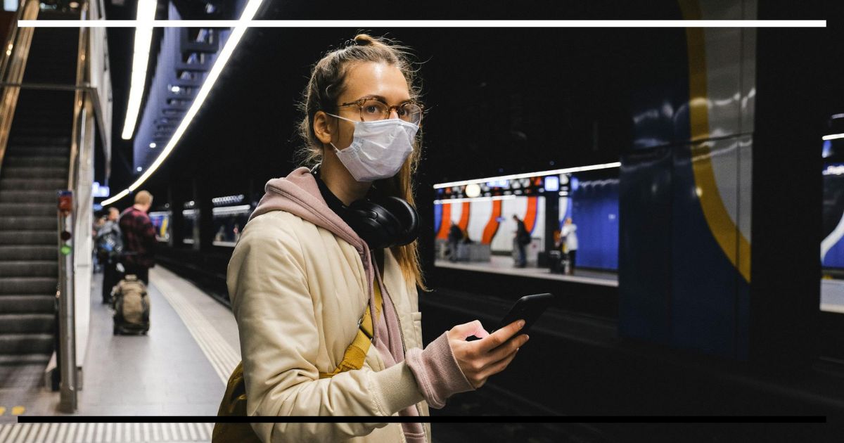 Jovem com máscara e celular em estação de metrô, cercada por telas e luzes artificiais.