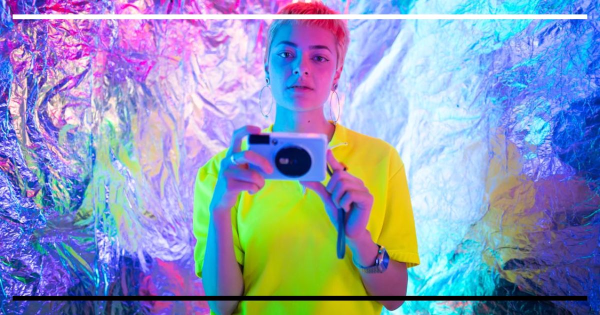 Jovem com cabelo curto e camisa fluorescente segura uma câmera diante de fundo metálico colorido em estilo Y2K.
