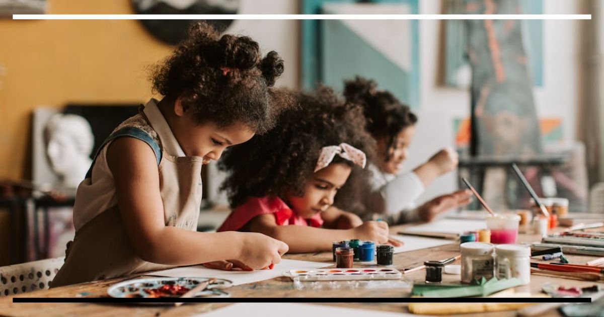 Crianças pintando juntas em uma mesa de arte, concentradas na atividade, representando como o ambiente estimula o aprendizado e o desenvolvimento infantil.