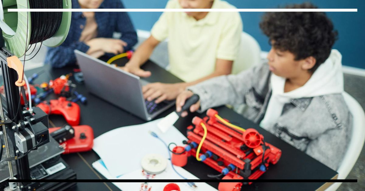 Três crianças programando em um laptop, com uma impressora 3D e robôs em uma aula de Alfabetização Digital.