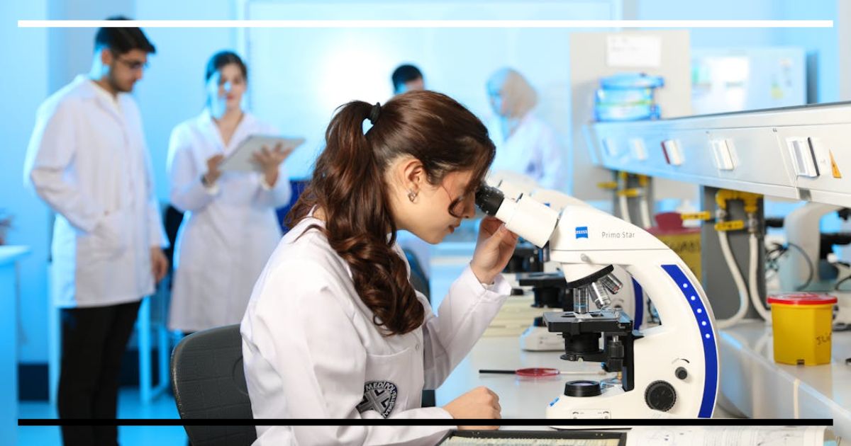 Estudante observando amostras em um microscópio em laboratório, com outros alunos ao fundo.