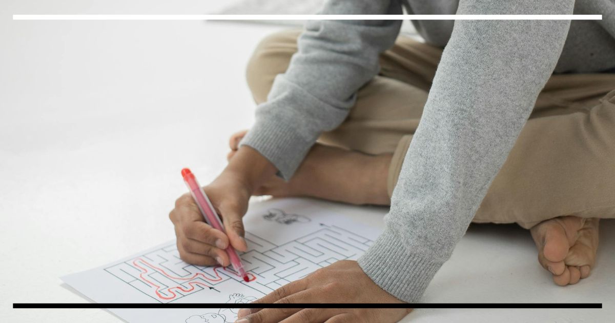Criança resolvendo labirinto com lápis vermelho, pensando logicamente no chão branco.