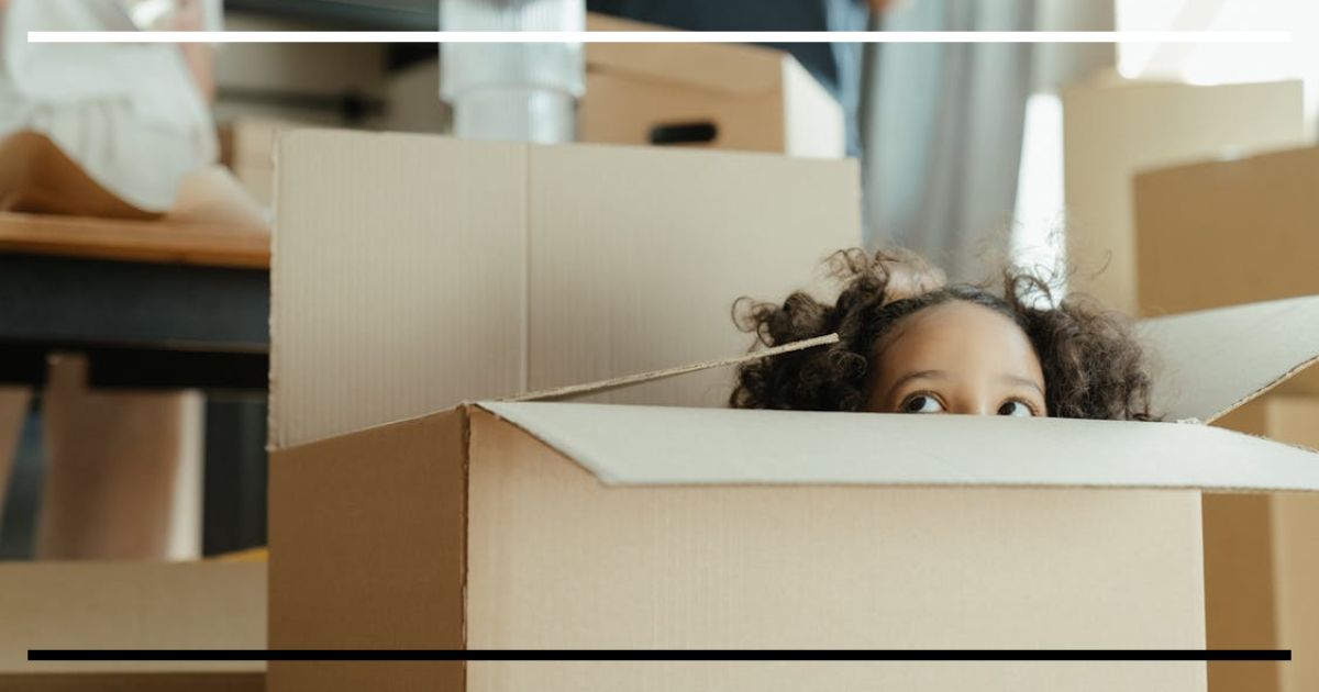 Criança de cabelo cacheado espiando curiosa de dentro de uma grande caixa de papelão, ilustrando a criatividade low-cost.