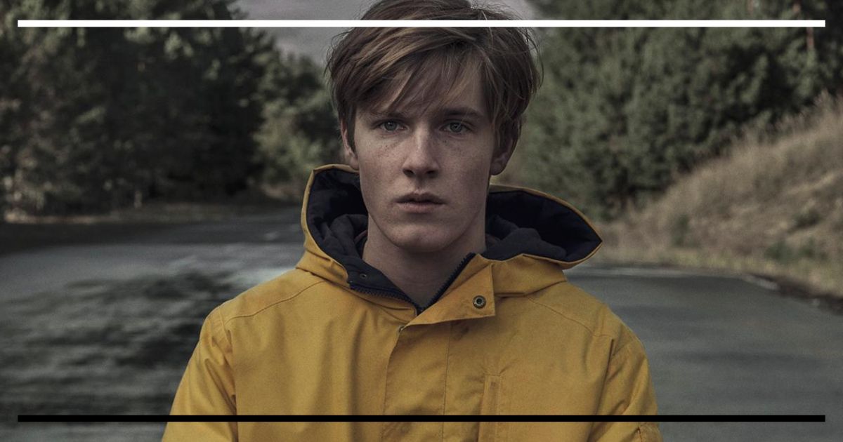 Close-up frontal do ator Louis Hofmann no papel de Jonas Kahnwald na série Dark (Netflix), vestindo sua icônica capa de chuva amarela, com uma expressão séria. Ao fundo, uma estrada e árvores cinzentas da floresta de Winden.