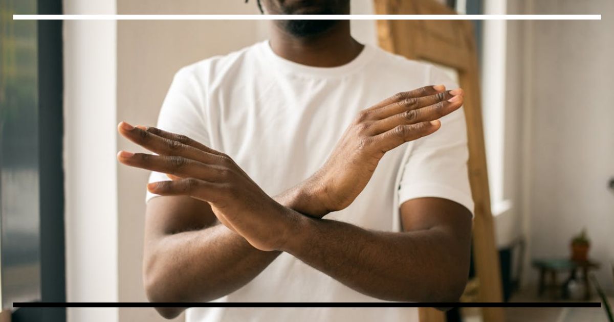 Homem com camiseta branca faz gesto de “não” com os braços cruzados em X diante do corpo.