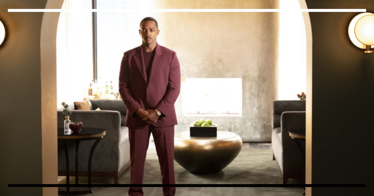 Texto alternativo: Anthony Mackie (Tom) em pé, centralizado em uma sala de estar moderna e luxuosa. Ele veste um terno vinho impecável e tem uma expressão séria e estoica, olhando diretamente para a câmera.