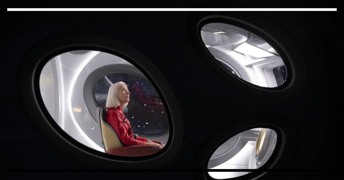 Texto alternativo: Helen Mirren (Peg) vestindo um traje espacial vermelho, sentada de perfil dentro de uma nave escura. Ela olha para cima através de três janelas ovais que mostram o espaço sideral.