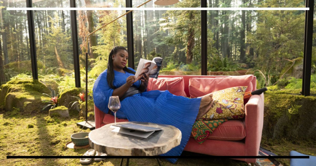 Uzo Aduba (Sasha) deitada confortavelmente em um sofá rosa, lendo um livro e segurando uma taça de vinho. Ela está em uma sala com paredes inteiramente de vidro, cercada por uma floresta densa do lado de fora.