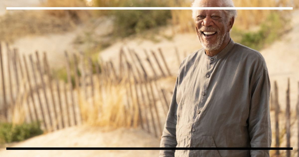 Morgan Freeman (Stuart) em plano médio, vestindo roupas cinzas simples, rindo abertamente em uma praia ensolarada com cercas de madeira ao fundo.