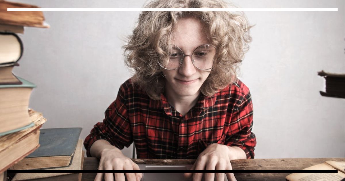 Jovem com óculos digitando em notebook rodeado de livros antigos em mesa de madeira