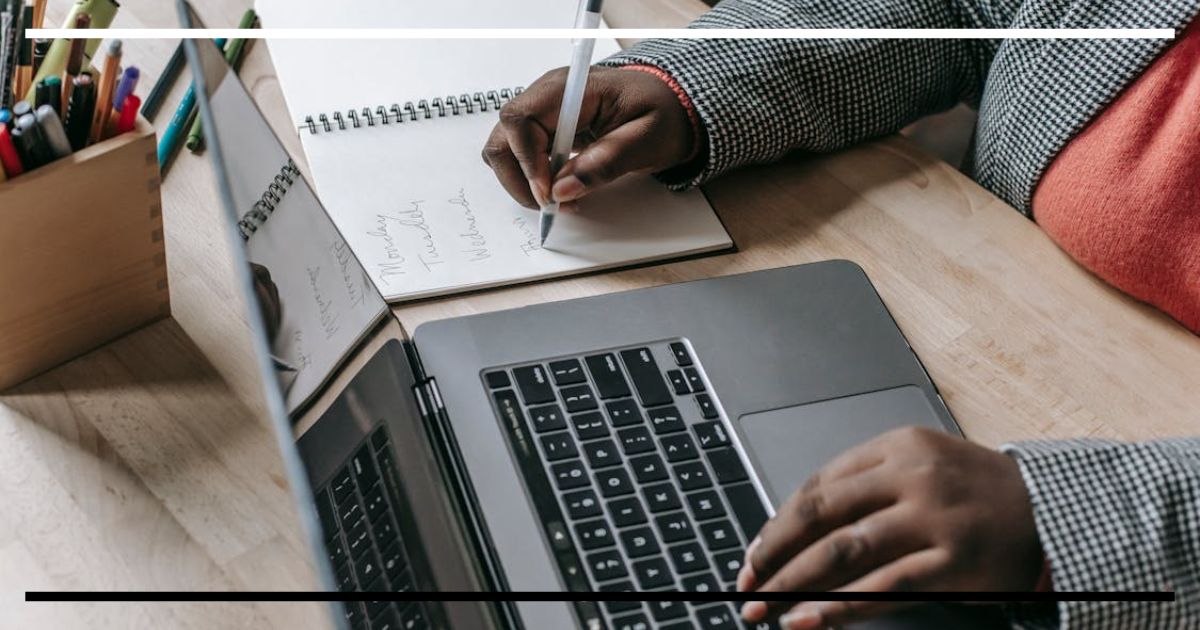 Pessoa escrevendo em caderno com caneta enquanto usa notebook em mesa de estudo.