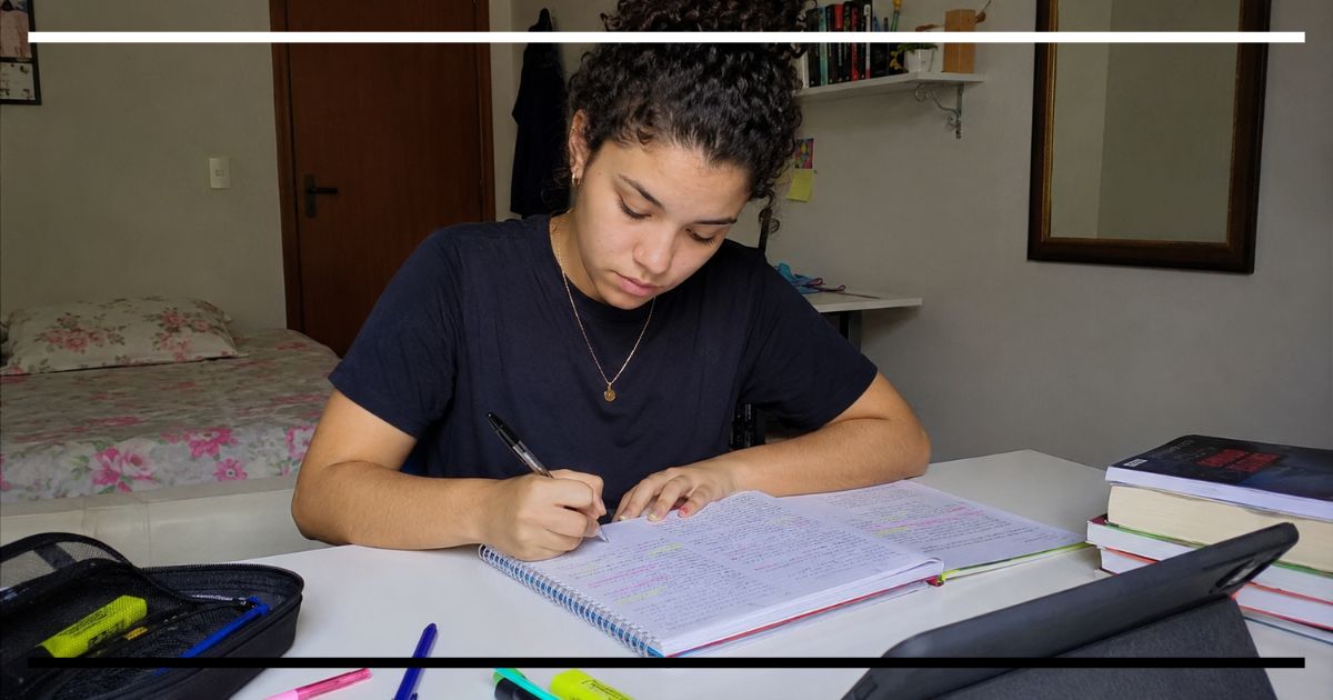 Estudante escrevendo um resumo em caderno aberto, com livros, canetas e material de estudo sobre a mesa.