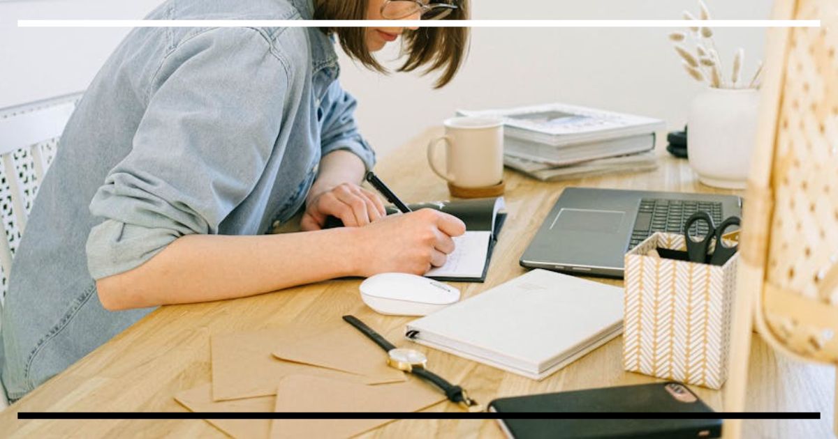 Pessoa estudando à mesa, escrevendo em um caderno com dispositivo digital ao lado.