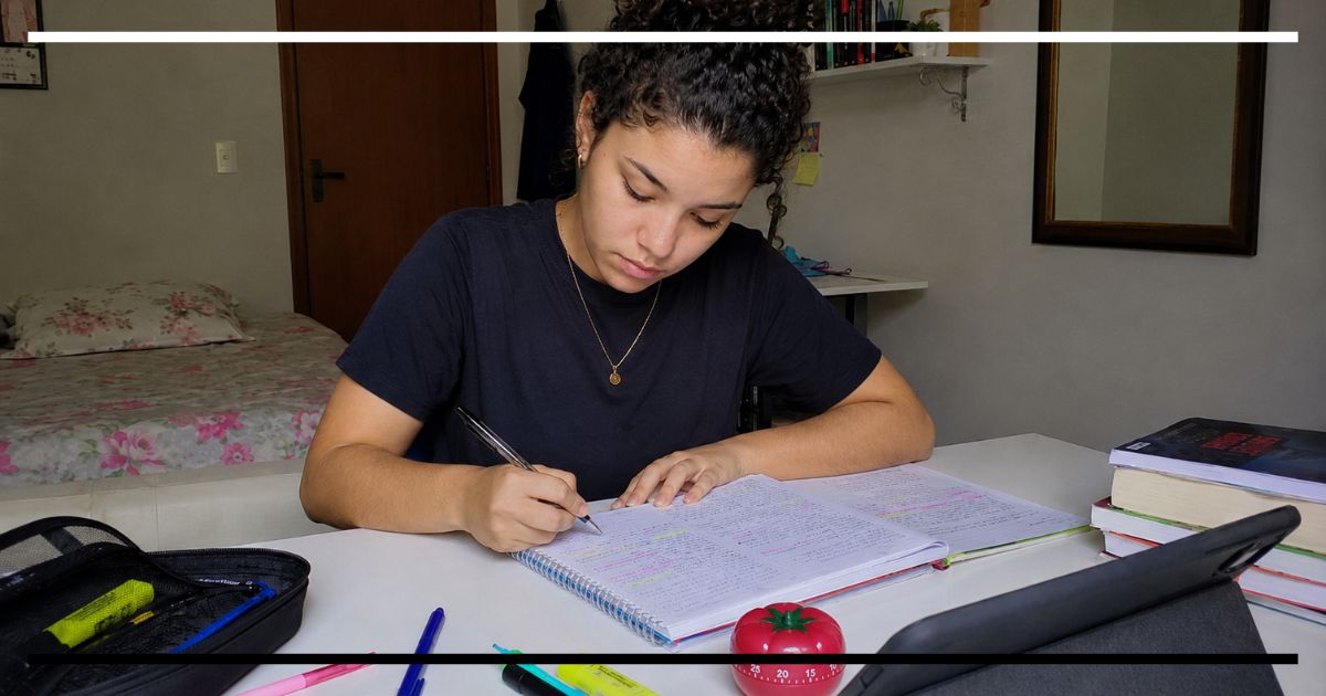 Estudante escrevendo em caderno com um timer Pomodoro sobre a mesa de estudos.