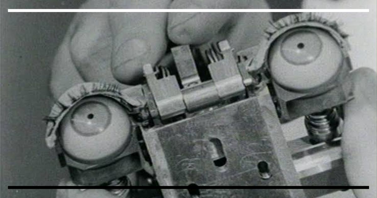 Fotografia em preto e branco de uma mão segurando um mecanismo mecânico com dois olhos artificiais de autômato, com engrenagens e molas visíveis.