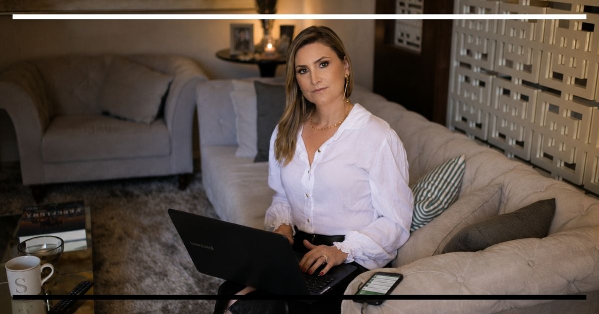 Mulher trabalha com notebook no sofá de casa, com celular e materiais de trabalho ao redor, representando a sobreposição entre vida profissional e pessoal.